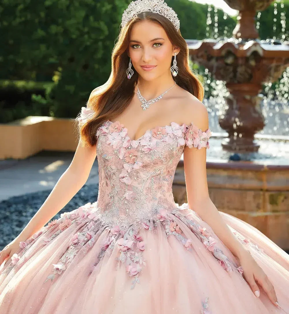 Joven con vestido de quinceañera rosa bordado, corona y joyería, junto a una fuente elegante.