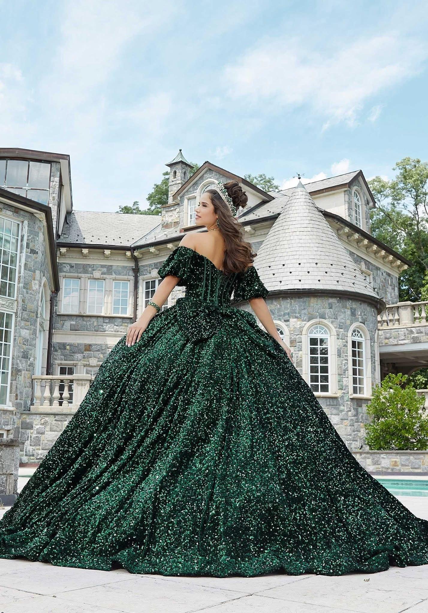 Joven con vestido de quinceañera verde esmeralda brillante y corona, posando frente a mansión elegante.