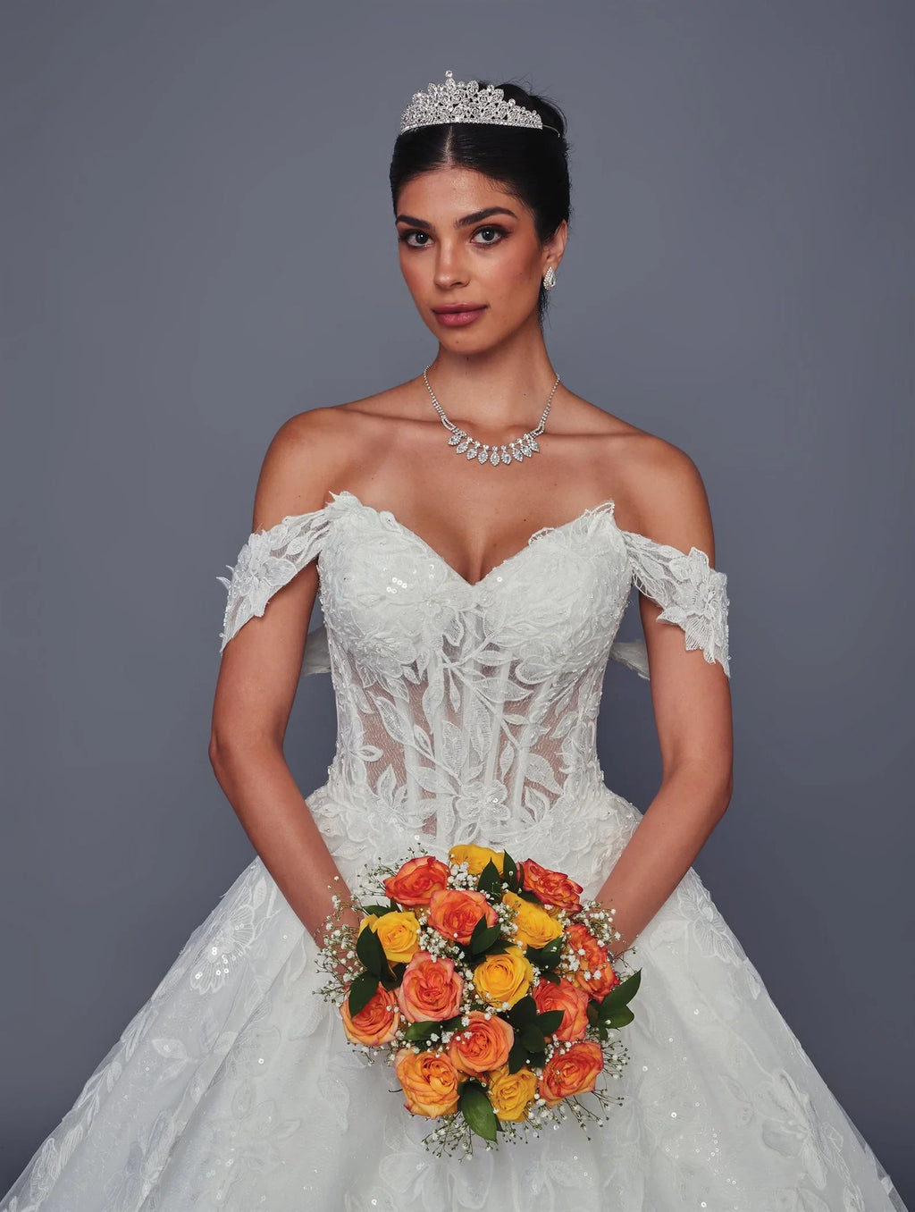 Novia con vestido de encaje línea A de DeKlaire Bridal, tiara y ramo de rosas naranjas.