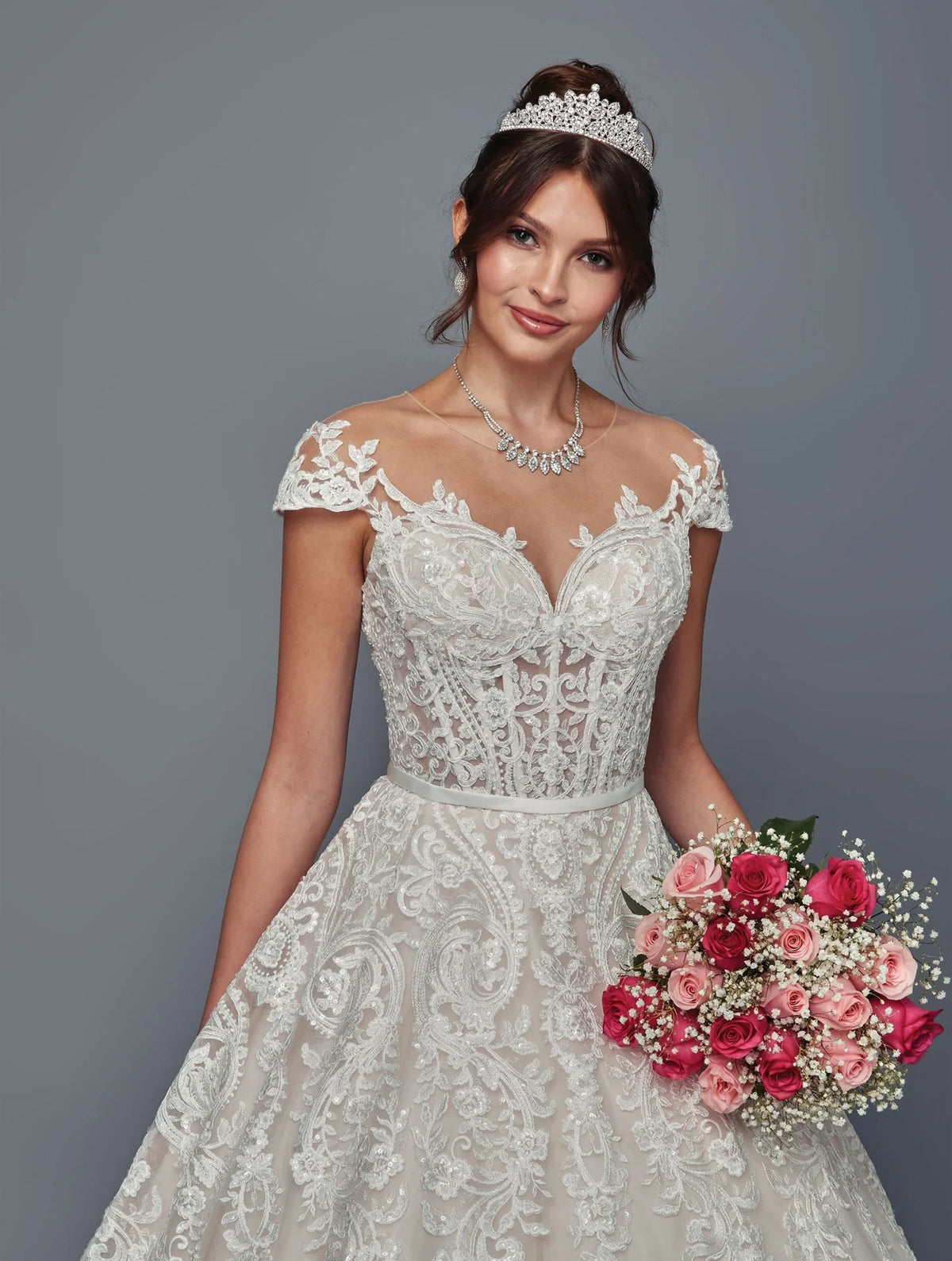 Novia con vestido de línea A DeKlaire Bridal, escote ilusión, encaje y ramo de rosas.