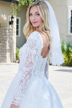 Novia con vestido de encaje blanco, manga larga y velo, en exterior soleado.