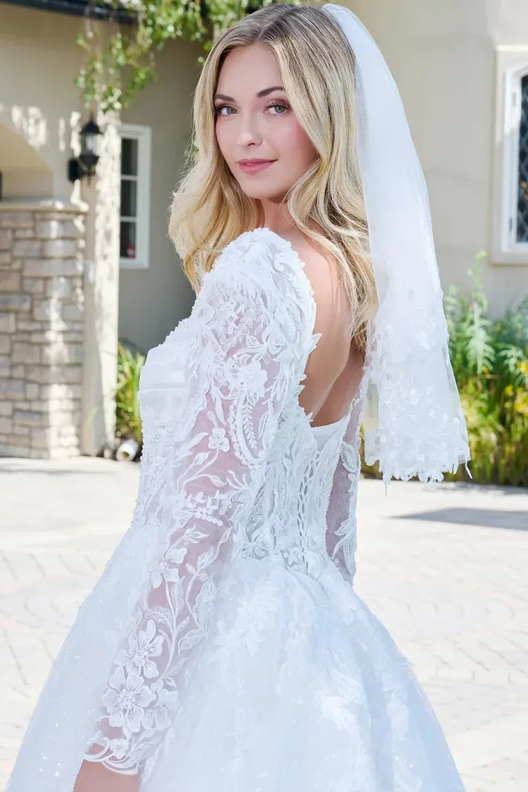 Novia con vestido de encaje blanco, manga larga y velo, en exterior soleado.