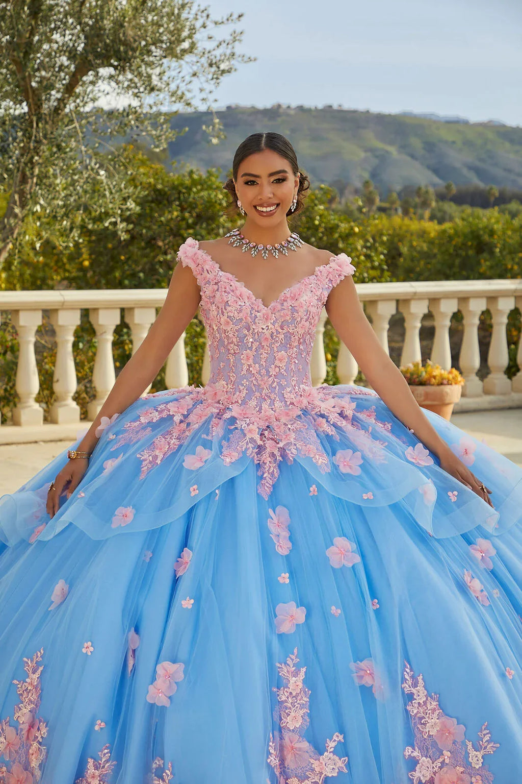 Joven con vestido de quinceañera azul y rosa, detalles florales, en jardín elegante