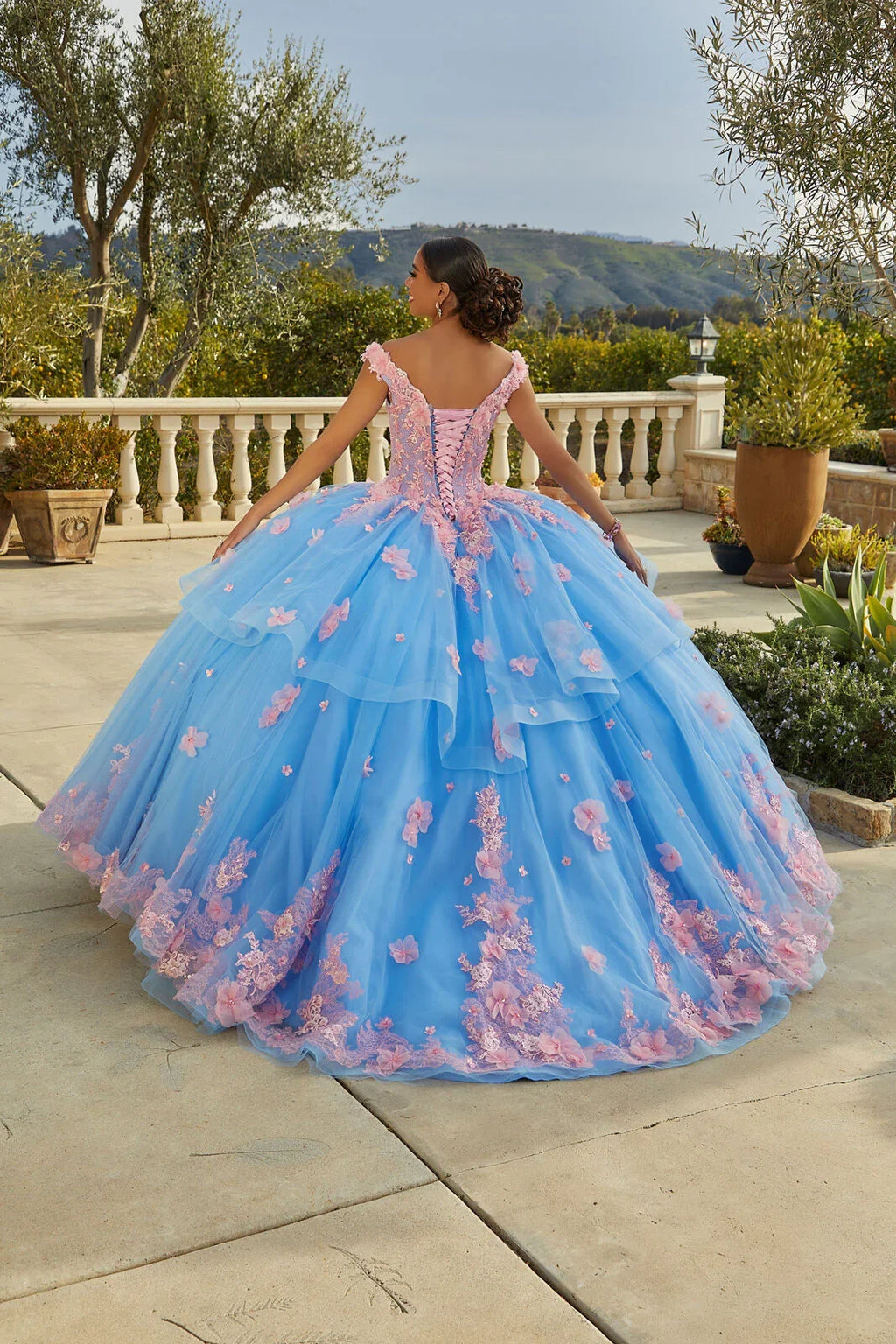 Joven de espaldas con vestido de quinceañera azul con flores rosas en un jardín elegante